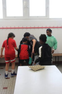 A group of children huddle around a table in a classroom or gym, with an egg carton on the white table in the foreground.
