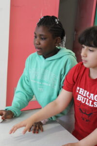 Two children lean over a light table: girl in a teal Cambridge hoodie and a boy in a red Chicago Bulls shirt reach toward the surface.