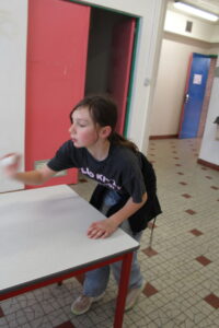 Girl leaning over a gray table in a school hallway, throwing a white ball toward the table edge.