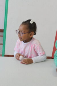 Young girl with glasses and a white hair clip sits at a table holding an egg, preparing to crack it, in a classroom setting.