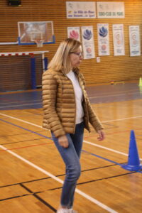 Woman in a tan puffer jacket and jeans walks across an indoor gym with a basketball hoop and banners on the wall nearby.