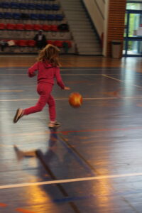 Young girl in a pink tracksuit runs across a gym floor toward a basketball in motion.