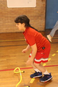 Young girl in a red sports uniform bends forward in a gym, with a yellow hula hoop on the wooden floor nearby.