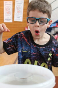 Boy wearing blue protective glasses and a colorful shirt, making an astonished face while reaching toward a shallow bowl of water on a table in a classroom.