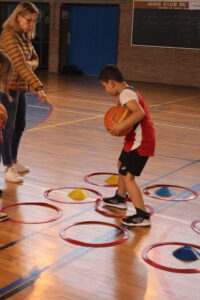Young boy in a red basketball uniform dribbles a ball through red hoops and yellow cones in a gym as a coach watches.