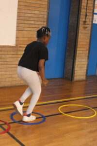 A person in a gym stepping through a blue hoop on the wooden floor, wearing a black shirt, light gray leggings, and white sneakers near brick walls.