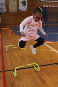 Girl in pink outfit jumping over a small yellow hurdle in a gymnasium floor.