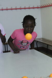 Girl in pink shirt leaning over a table, watching an orange ball mid‑air during a game.