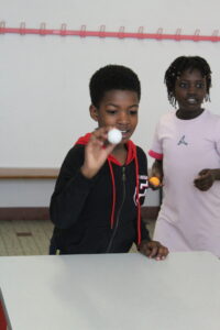 Boy in a black hoodie at a table holds up a small white ball toward the camera as a girl in pink looks on nearby in a room with a pink rack on the wall behind them.