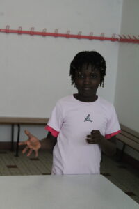 Girl standing indoors, wearing a light pink shirt with a logo, gesturing with her left hand extended toward the camera; pink hooks along the wall in the background.