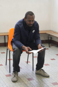 Man in a navy Nike jacket sits on an orange chair, examining a piece of paper in a tiled room.