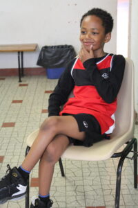Boy in red and black basketball uniform sits on a beige chair, resting chin on hand with a thoughtful smile in a gym-like room.
