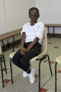 A young girl sits on a beige chair in a room, wearing a white blouse, black pants, white sneakers, and polka-dotted socks, hands folded in her lap.