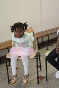 Young girl sitting on a chair in a light-colored room, wearing a pastel mint top, pink skirt, white tights, and gold sneakers, with one hand raised behind her.