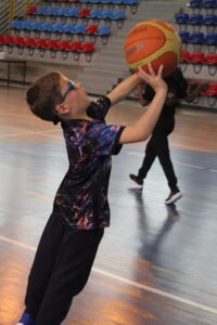 Boy in a patterned shirt and blue glasses reaches to catch a orange basketball in a gym.