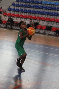 Young basketball player in a green uniform prepares to shoot a ball inside an indoor gym with blue and red spectator seating in the background