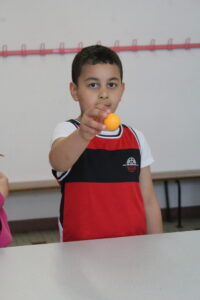 Young boy in a red and black jersey holds an orange ball toward the camera in a classroom setting.
