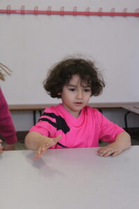 Child in a bright pink shirt reaches forward with her hand while seated at a table in a classroom setting.