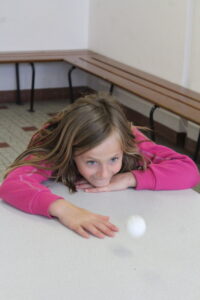 Girl in a pink hoodie leaning on a table, reaching toward a small white ball on the surface.