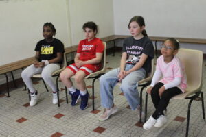 Four children sit in a row on plastic chairs in a room, watching something off-camera.