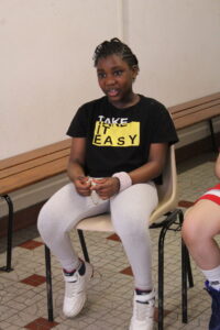 Young girl with braided hair, wearing a black 'TAKE IT EASY' shirt, sits on a chair indoors and holds a bracelet.