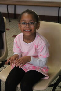 Smiling young girl with glasses in a pink shirt, seated on a beige chair indoors.
