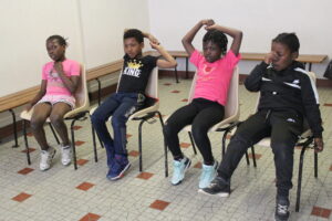 Four children sit in a row on plastic chairs, stretching their arms overhead in a tiled indoor space.