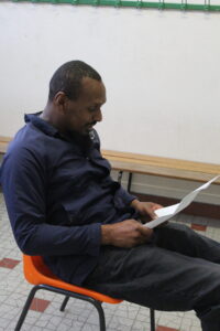Man with short hair and beard seated in an orange chair, reading a sheet of paper.