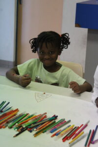 Smiling girl with braided hair drawing a pizza slice at a table with colorful pencils laid out nearby.
