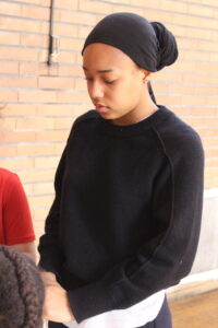 Person with a black head wrap and black sweater, looking down beside a brick wall.