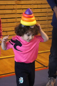 Child in a pink shirt balances a rainbow-colored ring stack on their head in a gym.