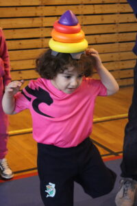 Child in a pink shirt balances a colorful stacking ring toy on their head in a gym.