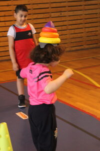 Child wearing a hat made of stacked colorful plastic rings during indoor gym activity, with another child watching in the background.