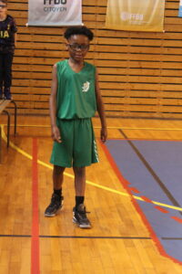 Young boy in a green basketball uniform standing on a gym floor with wooden bleachers in the background.