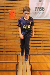 Young boy with blue glasses balancing on a gym beam in a gymnasium, wall has an orange poster with a spider.