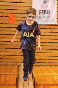 Boy with blue glasses balances on a wooden gym bench, wearing a colorful shirt with the letters AIA.