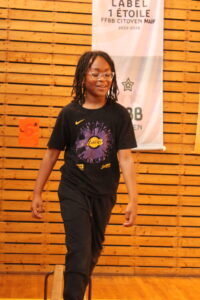 Young person with glasses and dreadlocks walking in a gym, wearing a Los Angeles Lakers t‑shirt, with a wooden wall and banner in the background.