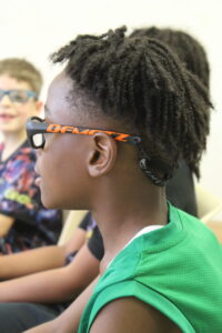 Side profile of a boy wearing black-framed glasses with orange temples, in a green sports jersey, indoors.