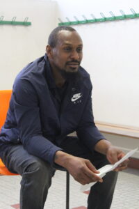 Man with short hair and goatee sitting on an orange chair, reading papers in a bright room in a navy Nike jacket.