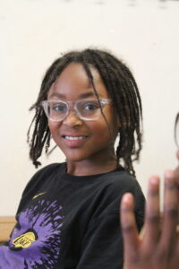 Smiling girl with clear-framed glasses and braided hair, wearing a black Lakers T‑shirt, reaching toward the camera.
