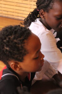 Side profile of a young woman in a white shirt seated beside a child with short curly hair indoors, both looking downward.