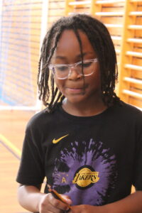 Child with braids in clear glasses, wearing a black Lakers T-shirt, holding a pencil in a gym setting with wooden bleachers behind him/her.