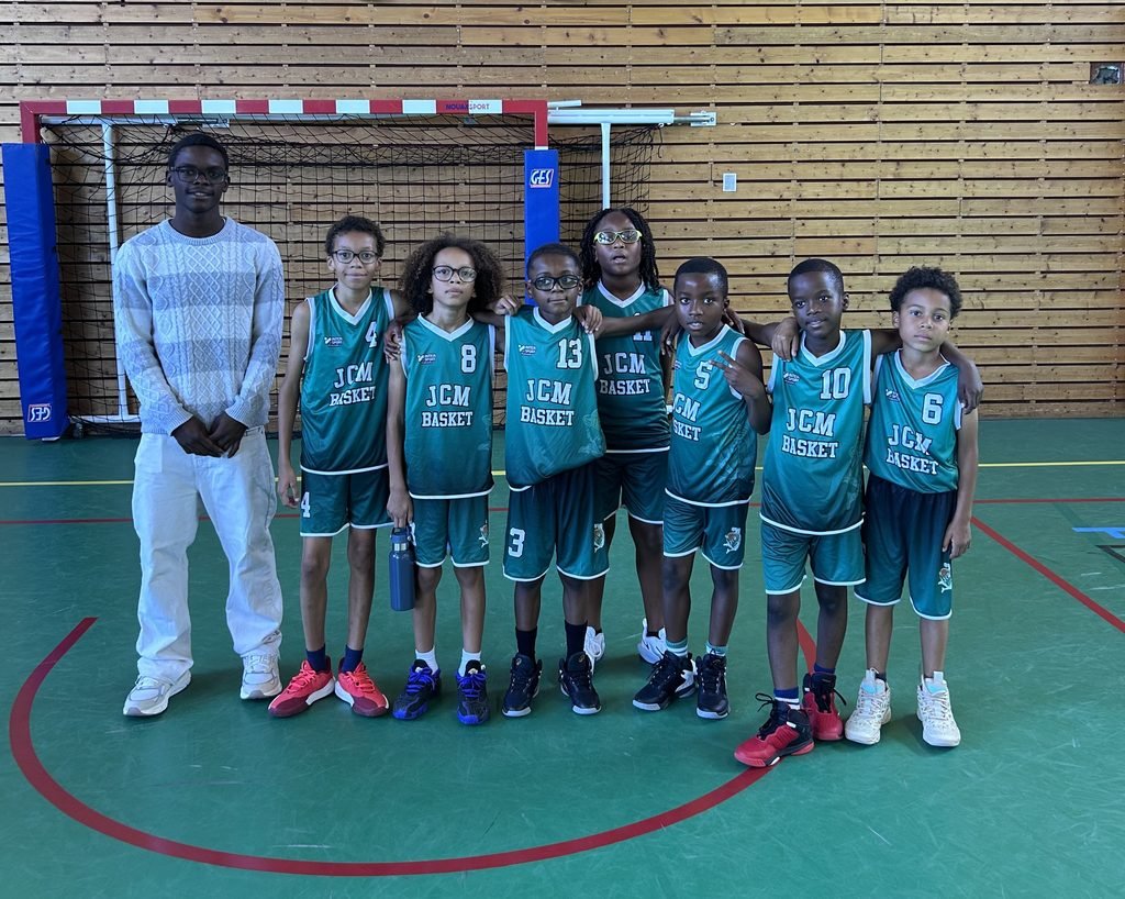 Group of young basketball players in teal uniforms standing with a tall man in a light sweater inside a gym, arms around shoulders, posing for a team photo.