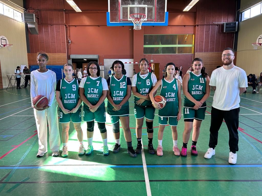 Youth girls' basketball team posing for a photo in an indoor gym with a coach on the right and a player holding a ball on the far left